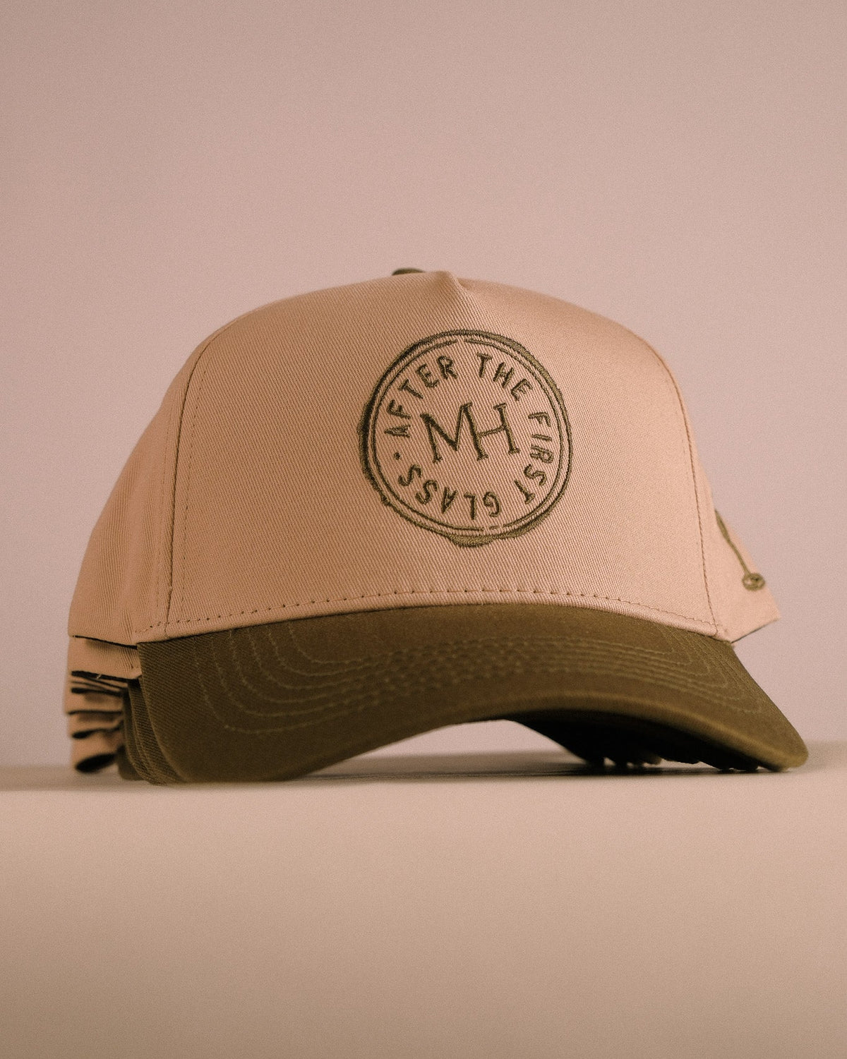 A beige and olive green trucker cap with a circular embroidered logo on the front, displayed against a solid beige background.
